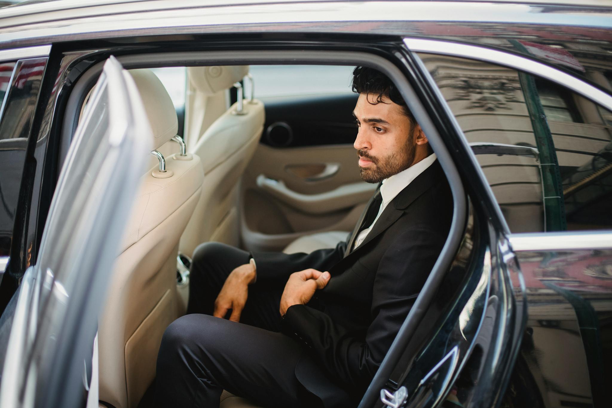A bearded man in a black suit sits elegantly inside a luxury car, exuding sophistication.