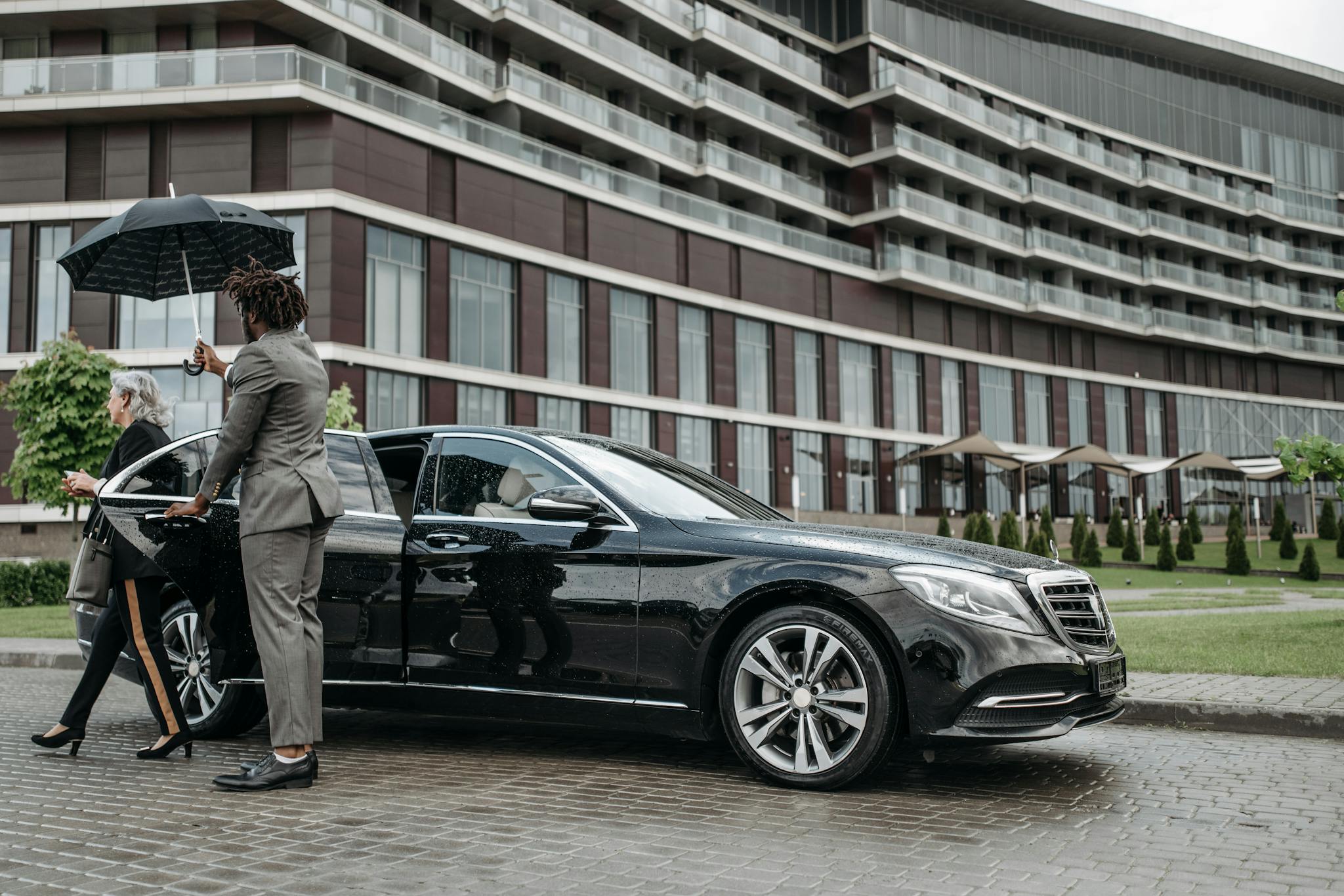 A business professional exits a luxury car while being shielded by an umbrella.
